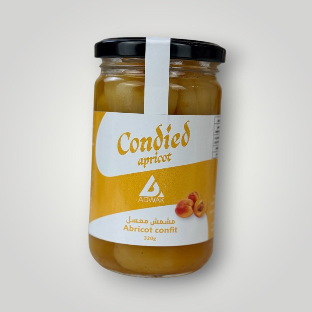 Bocal en verre cylindrique de 320 g d'abricots confits, couvercle métallique noir avec banderole blanche. Étiquette dorée à fond jaune intense, inscription "Condied apricot – Abricot confit 320g" en arabe et français avec illustration d'abricots frais orange. Le contenu — morceaux d'abricots confits dorés baignant dans leur sirop — est visible à travers le verre. Fond blanc uni.