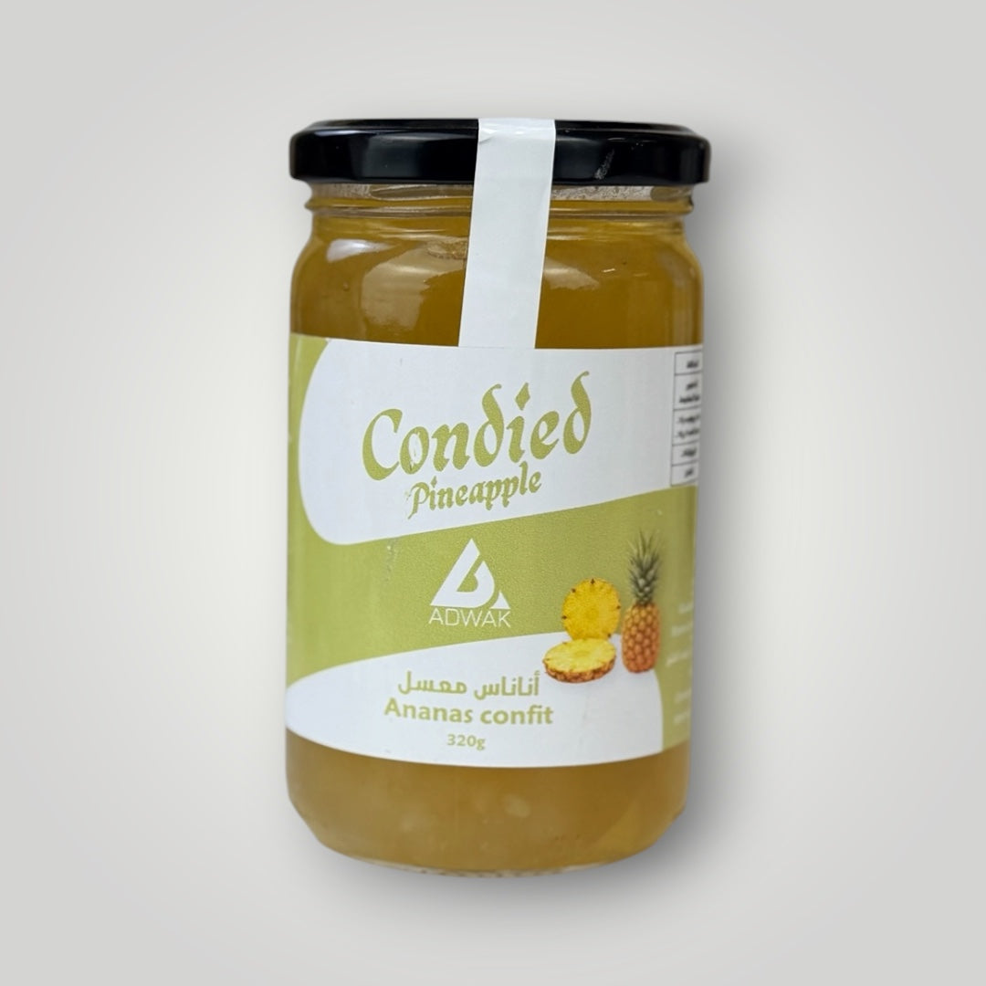 Bocal en verre cylindrique de 320 g d'ananas confit, couvercle métallique noir avec banderole blanche. Étiquette fond vert-jaune clair, inscription "Condied Pineapple – Ananas confit 320g" en arabe et français avec illustration d'un ananas entier et de tranches. Le contenu doré-ambré — morceaux d'ananas confits dans leur sirop épais — est visible à travers le verre. Fond gris clair dégradé.
