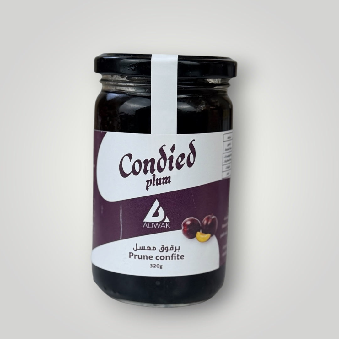 Bocal en verre cylindrique de 320 g de prunes confites, couvercle métallique noir avec banderole blanche. Étiquette fond violet foncé, inscription "Condied plum – Prune confite 320g" en arabe et français avec illustration de prunes violettes et chair jaune. Le contenu très sombre — prunes confites presque noires dans un sirop dense — est visible à travers le verre. Fond gris clair