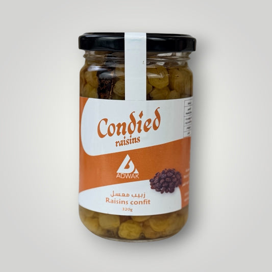 Bocal en verre cylindrique de 320 g de raisins confits, couvercle métallique noir avec banderole blanche. Étiquette fond orange vif, inscription "Condied raisins – Raisins confit 320g" en arabe et français avec illustration de raisins secs. Les grains de raisins confits vert-doré, bien gonflés et brillants, sont clairement visibles à travers le verre. Fond blanc-gris uni.