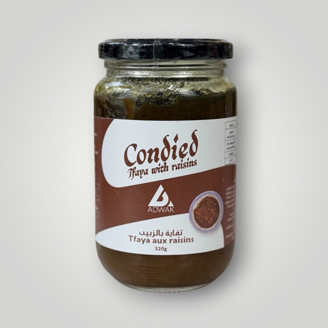 Bocal en verre cylindrique de 320 g de tfaya aux raisins, couvercle métallique noir avec banderole blanche. Étiquette fond terracotta-brun, inscription "Condied Tfaya with raisins – Tfaya aux raisins 320g" en arabe et français, avec illustration d'un bol de sauce marocaine. Le contenu brun-vert foncé — oignons caramélisés confits aux raisins — est visible en surface du bocal non entièrement rempli. Fond blanc-gris clair.