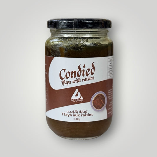 Bocal en verre cylindrique de 320 g de tfaya aux raisins, couvercle métallique noir avec banderole blanche. Étiquette fond terracotta-brun, inscription "Condied Tfaya with raisins – Tfaya aux raisins 320g" en arabe et français, avec illustration d'un bol de sauce marocaine. Le contenu brun-vert foncé — oignons caramélisés confits aux raisins — est visible en surface du bocal non entièrement rempli. Fond blanc-gris clair.